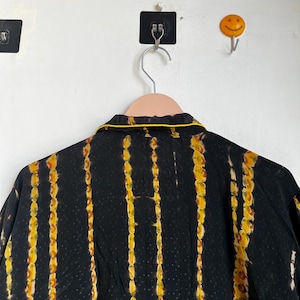 Może przedstawiać: Czarna koszula z pionowymi ż&oacute;łto-pomarańczowymi paskami tie-dye. Koszula ma ż&oacute;łte wykończenie wok&oacute;ł kołnierza. Koszula wisi na r&oacute;żowym wieszaku na białej ścianie.