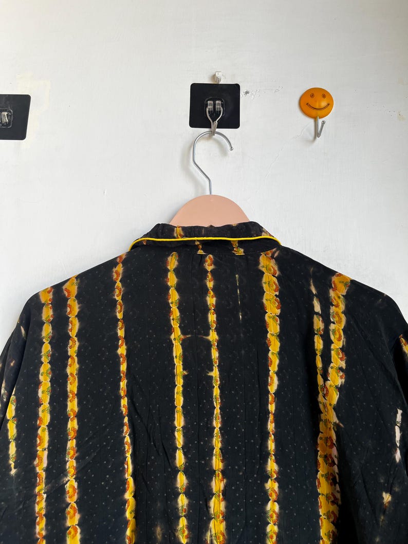 Może przedstawiać: Czarna koszula z pionowymi ż&oacute;łto-pomarańczowymi paskami tie-dye. Koszula ma ż&oacute;łte wykończenie wok&oacute;ł kołnierza. Koszula wisi na r&oacute;żowym wieszaku na białej ścianie.