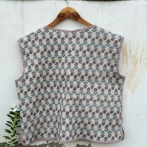 May include: A sleeveless, quilted vest with a floral pattern in blue and gray on a beige background. The vest has a scalloped edge and is trimmed with a striped border. It is hanging on a wooden hanger.
