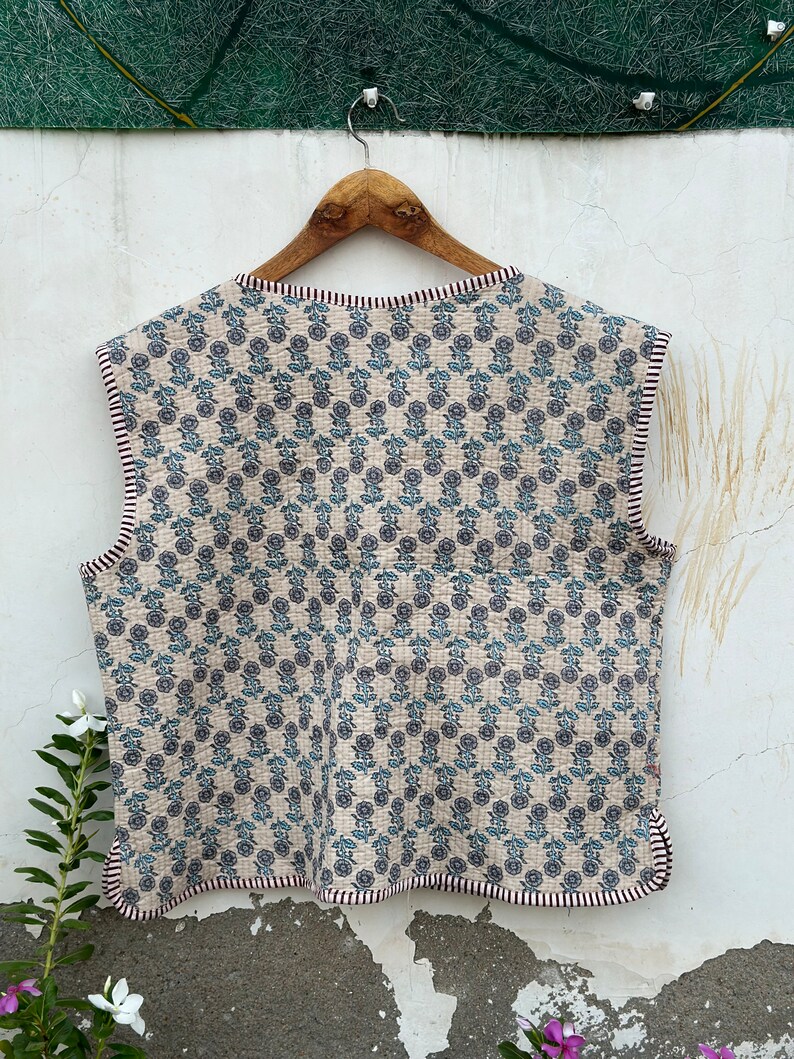 May include: A sleeveless, quilted vest with a floral pattern in blue and gray on a beige background. The vest has a scalloped edge and is trimmed with a striped border. It is hanging on a wooden hanger.