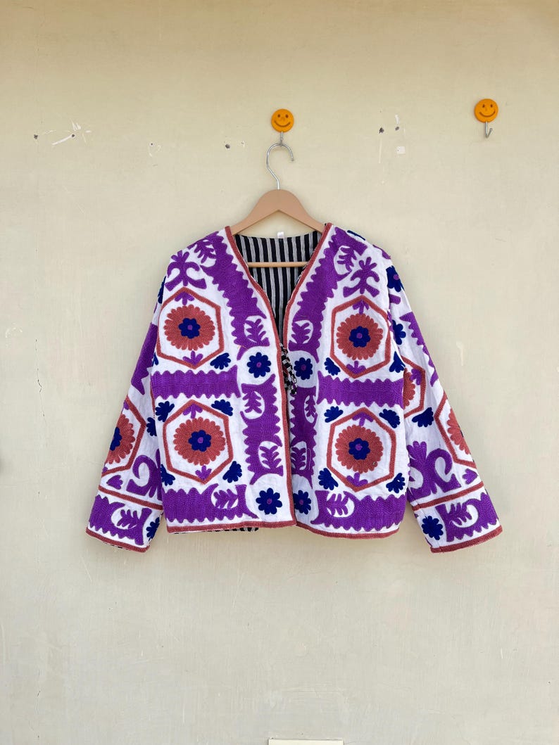 May include: A white jacket with purple and blue embroidery featuring a geometric floral pattern. The jacket has a V-neck and long sleeves.