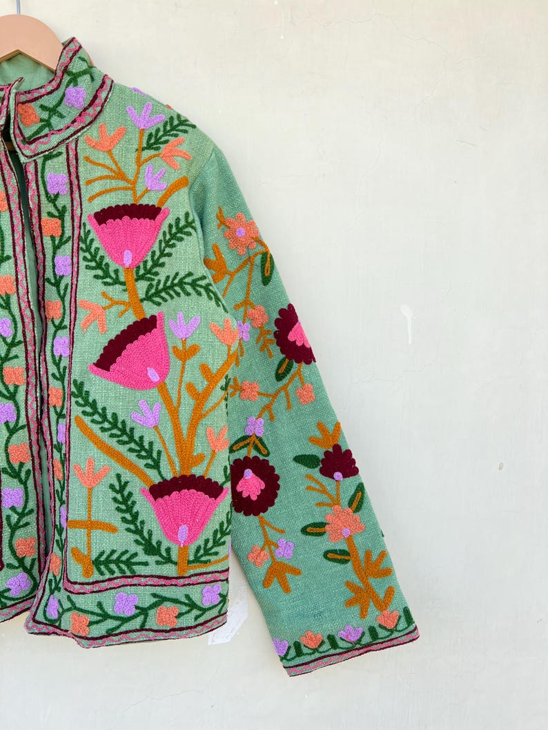 May include: A light green jacket with embroidered floral designs in shades of pink, orange, and purple. The jacket has a mandarin collar and a zipper closure.