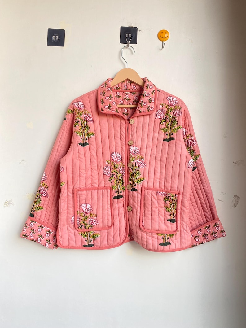 Pu&ograve; includere: Giacca trapuntata color corallo con stampa floreale. La giacca presenta un colletto, due tasche frontali e bottoni in legno. Il motivo floreale include fiori rosa e foglie verdi.