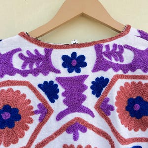 May include: White cotton top with colorful embroidered floral and geometric patterns in shades of purple, blue, and pink.