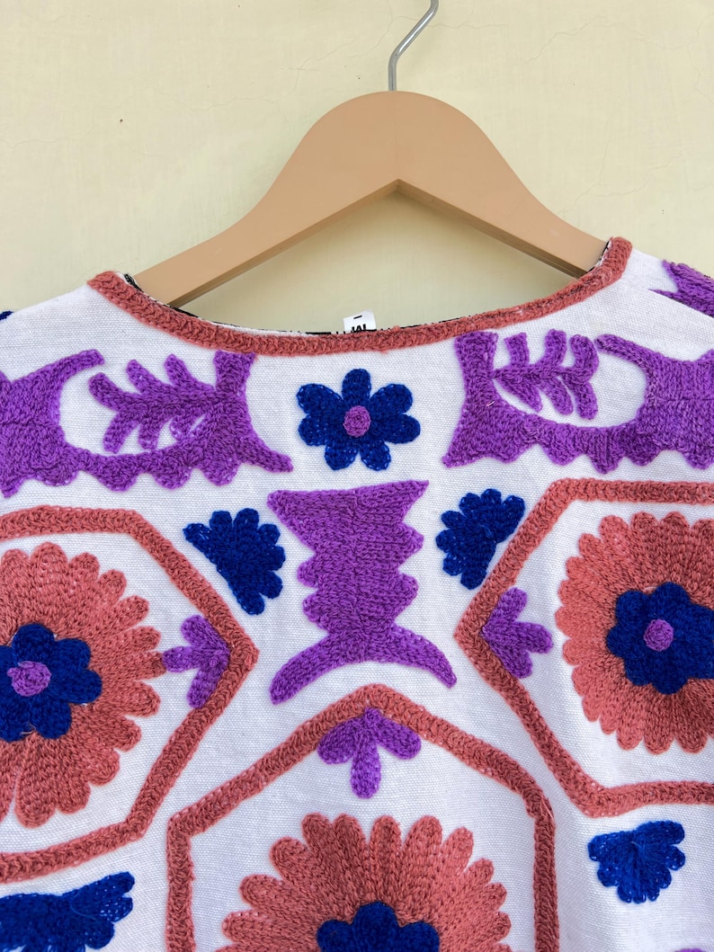 May include: White cotton top with colorful embroidered floral and geometric patterns in shades of purple, blue, and pink.