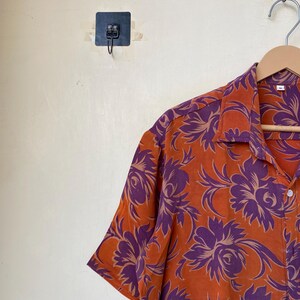 May include: Orange short-sleeve shirt with a purple floral pattern. The shirt is hanging on a wooden hanger. The shirt has a button-down front and a collar. The floral design is a repeating pattern.