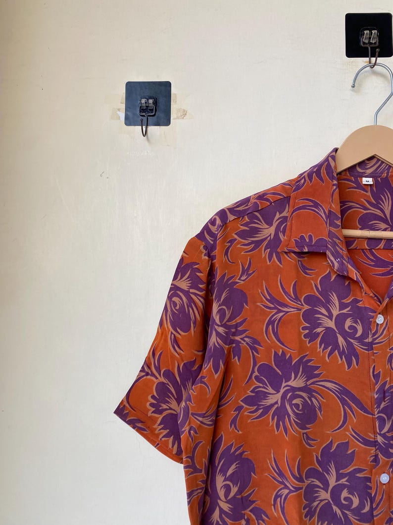May include: Orange short-sleeve shirt with a purple floral pattern. The shirt is hanging on a wooden hanger. The shirt has a button-down front and a collar. The floral design is a repeating pattern.