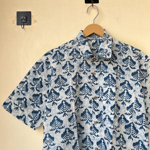May include: A short-sleeved button-down shirt with a blue and white leaf pattern. The shirt is hanging on a wooden hanger. The shirt has wooden buttons and a collared neckline. The background is a light beige color.