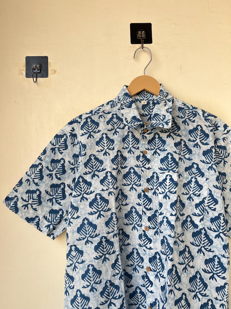 May include: A short-sleeved button-down shirt with a blue and white leaf pattern. The shirt is hanging on a wooden hanger. The shirt has wooden buttons and a collared neckline. The background is a light beige color.