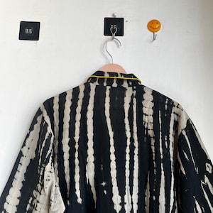 Może przedstawiać: Czarno-biała koszula w stylu tie-dye z pionowymi paskami, wisząca na r&oacute;żowym wieszaku. Koszula ma ż&oacute;łte wykończenie wok&oacute;ł kołnierza. Tło to biała ściana z czarnym kwadratowym haczykiem samoprzylepnym i ż&oacute;łtym haczykiem z uśmiechniętą buzią.