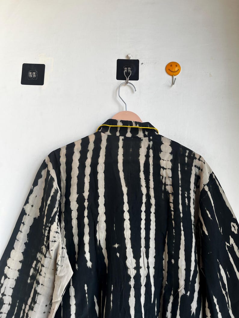 Może przedstawiać: Czarno-biała koszula w stylu tie-dye z pionowymi paskami, wisząca na r&oacute;żowym wieszaku. Koszula ma ż&oacute;łte wykończenie wok&oacute;ł kołnierza. Tło to biała ściana z czarnym kwadratowym haczykiem samoprzylepnym i ż&oacute;łtym haczykiem z uśmiechniętą buzią.
