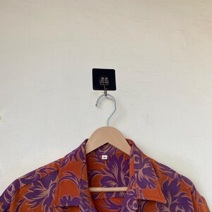 May include: An orange button-down shirt with a purple floral pattern hangs on a wooden hanger. The shirt is made of a lightweight fabric and has a classic collar. The background is a light beige wall.