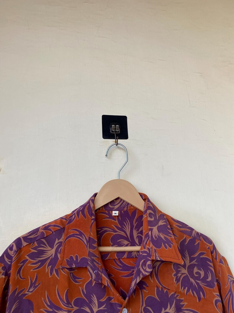 May include: An orange button-down shirt with a purple floral pattern hangs on a wooden hanger. The shirt is made of a lightweight fabric and has a classic collar. The background is a light beige wall.
