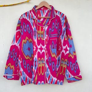May include: A pink, blue, and gold patterned long-sleeved shirt with a white trim. The shirt has a button-up front and a pocket on the left side.