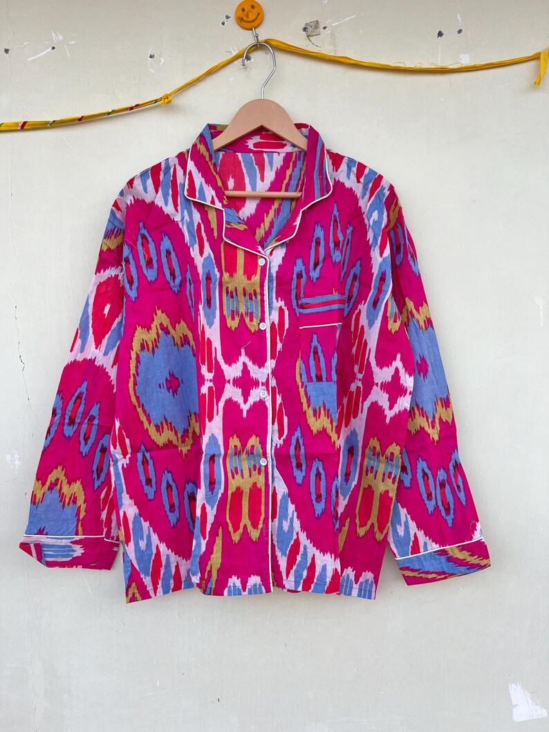 May include: A pink, blue, and gold patterned long-sleeved shirt with a white trim. The shirt has a button-up front and a pocket on the left side.