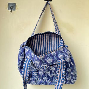 May include: A blue quilted tote bag with a floral pattern. The bag has a striped interior and matching striped handles. The bag is hanging on a wall. The floral pattern is white and light blue.