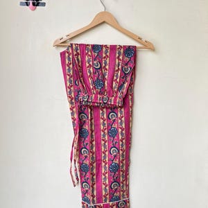 May include: Pink pajama set with vertical stripes and floral patterns in blue, cream, and red. The pajama pants and top are hanging on a wooden hanger against a white wall. The set features a drawstring waist and white trim.