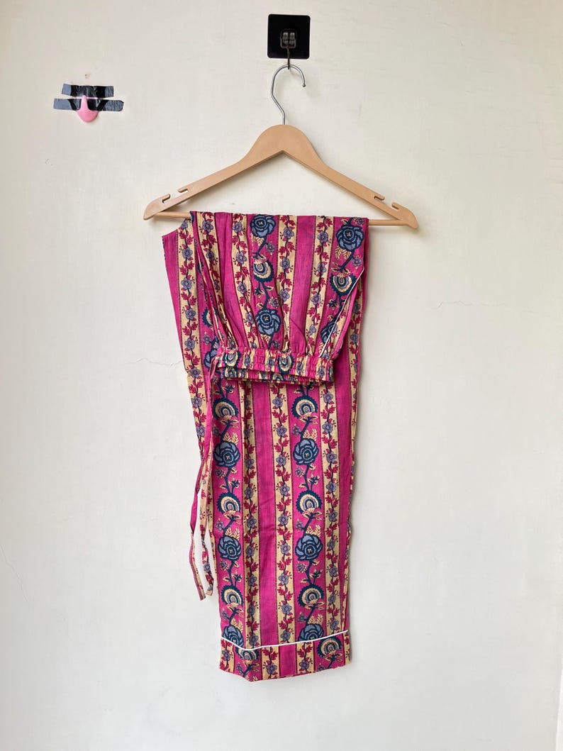 May include: Pink pajama set with vertical stripes and floral patterns in blue, cream, and red. The pajama pants and top are hanging on a wooden hanger against a white wall. The set features a drawstring waist and white trim.