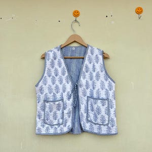 May include: A white cotton waistcoat with a blue and white striped pattern and a blue floral print. The waistcoat has two pockets and a tie closure.