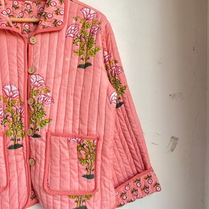 Pu&ograve; includere: Giacca trapuntata color corallo con stampa floreale. La giacca presenta un collo, bottoni in legno e due tasche anteriori. Il design floreale include fiori rosa e foglie verdi. La giacca &egrave; appesa a una gruccia di legno.
