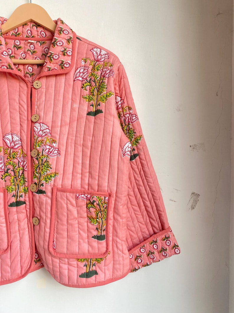 Pu&ograve; includere: Giacca trapuntata color corallo con stampa floreale. La giacca presenta un collo, bottoni in legno e due tasche anteriori. Il design floreale include fiori rosa e foglie verdi. La giacca &egrave; appesa a una gruccia di legno.