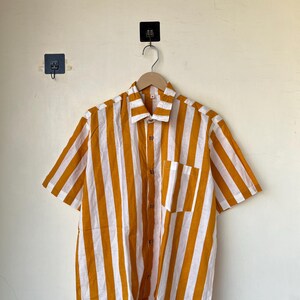 May include: Short-sleeved button-down shirt with vertical stripes in mustard yellow and white. The shirt has a chest pocket and wooden buttons. The shirt is hanging on a wooden hanger against a plain wall.