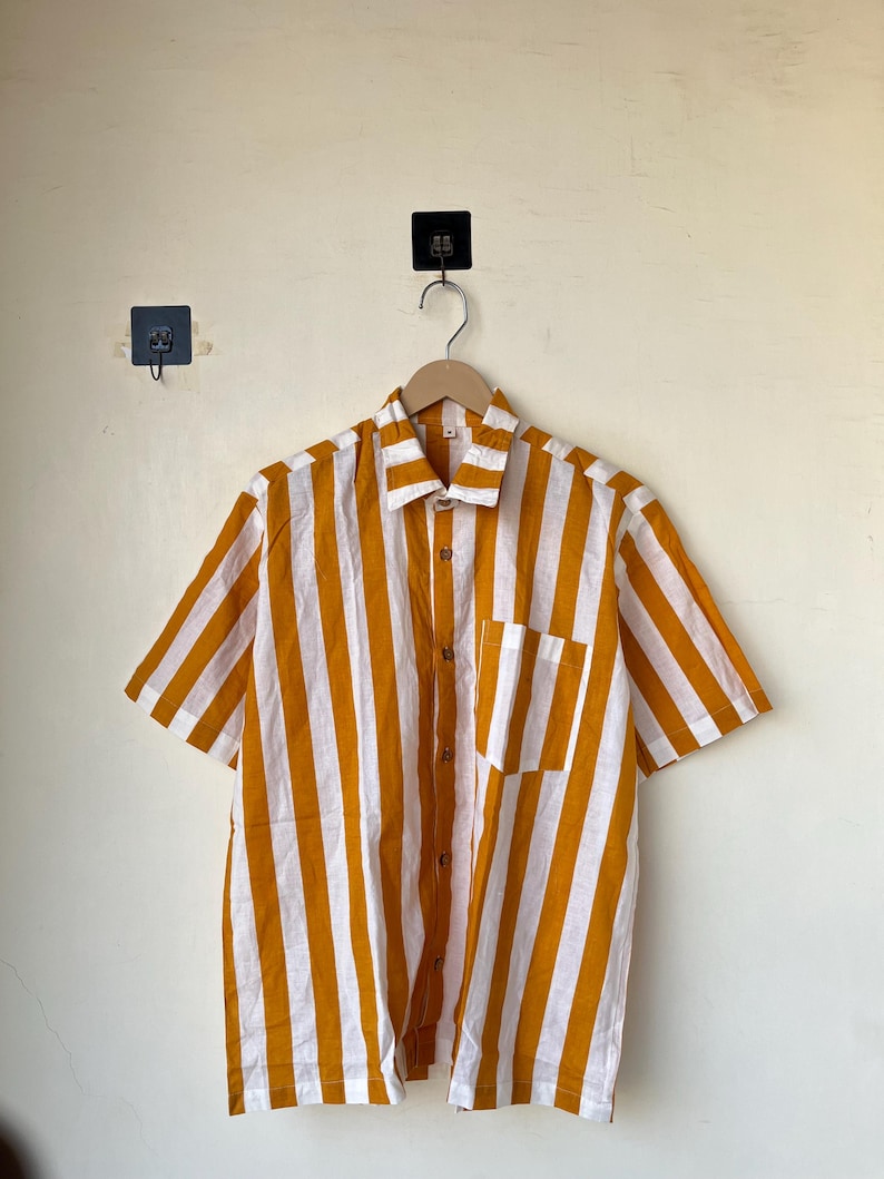 May include: Short-sleeved button-down shirt with vertical stripes in mustard yellow and white. The shirt has a chest pocket and wooden buttons. The shirt is hanging on a wooden hanger against a plain wall.