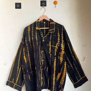 Może przedstawiać: Czarna koszula zapinana na guziki z długimi rękawami i ż&oacute;łto-czarnym wzorem tie-dye. Koszula ma ż&oacute;łte wykończenia wzdłuż kołnierza, mankiet&oacute;w i kieszeni. Koszula wisi na drewnianym wieszaku.
