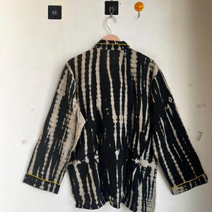 Może przedstawiać: Czarna i beżowa kurtka z efektem tie-dye z pionowymi paskami, wisząca na drewnianym wieszaku. Kurtka ma długie rękawy i ż&oacute;łte wykończenia wok&oacute;ł kołnierza i mankiet&oacute;w. Kurtka wisi na białej ścianie.