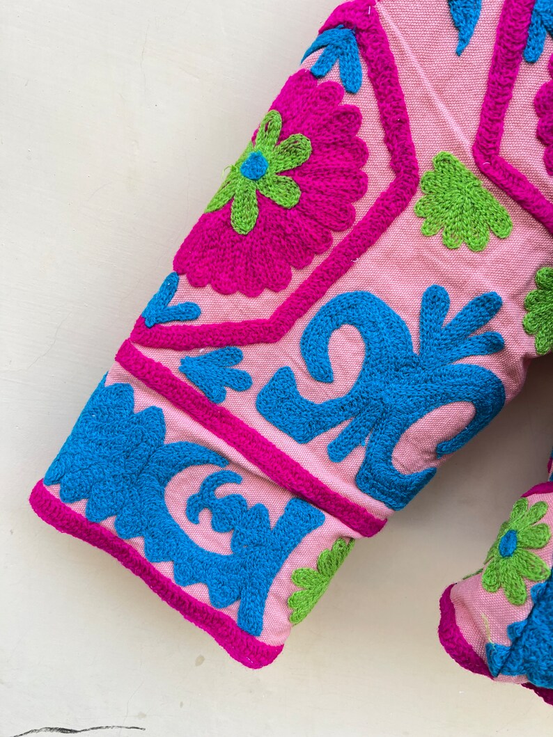 Puede incluir: Tela rosa con coloridos patrones bordados florales y geom&eacute;tricos en tonos rosa, azul y verde.