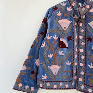 May include: A blue velvet jacket with floral embroidery in shades of pink, brown, and red. The jacket has a mandarin collar and a button closure.