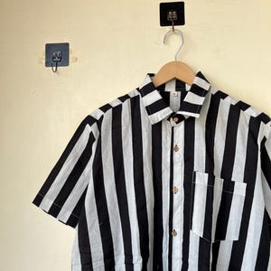 May include: Short-sleeved shirt with bold black and white vertical stripes. The shirt features a button-down front, a chest pocket, and wooden buttons. The garment is hanging on a wooden hanger.