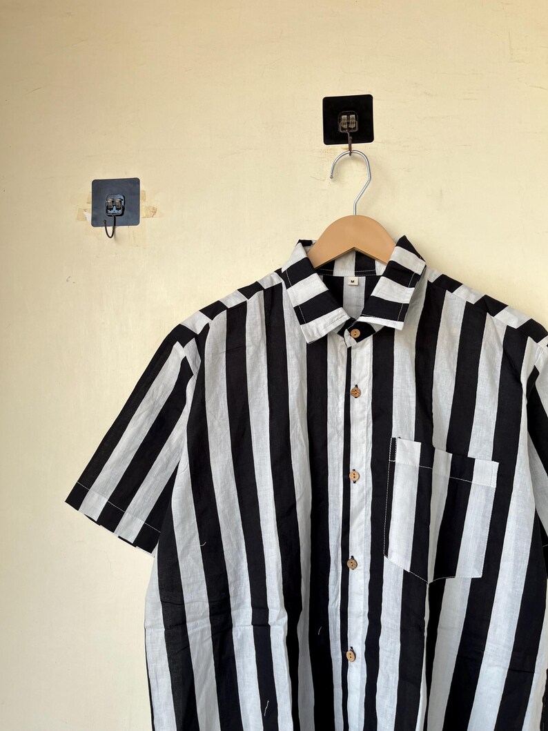 May include: Short-sleeved shirt with bold black and white vertical stripes. The shirt features a button-down front, a chest pocket, and wooden buttons. The garment is hanging on a wooden hanger.