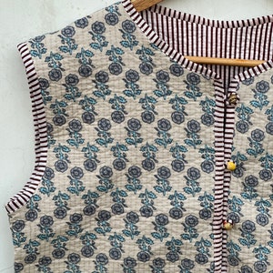 May include: A sleeveless, reversible quilted vest with a floral pattern in gray and light blue on a beige background. The vest has a striped border in white and burgundy, and is fastened with decorative buttons.