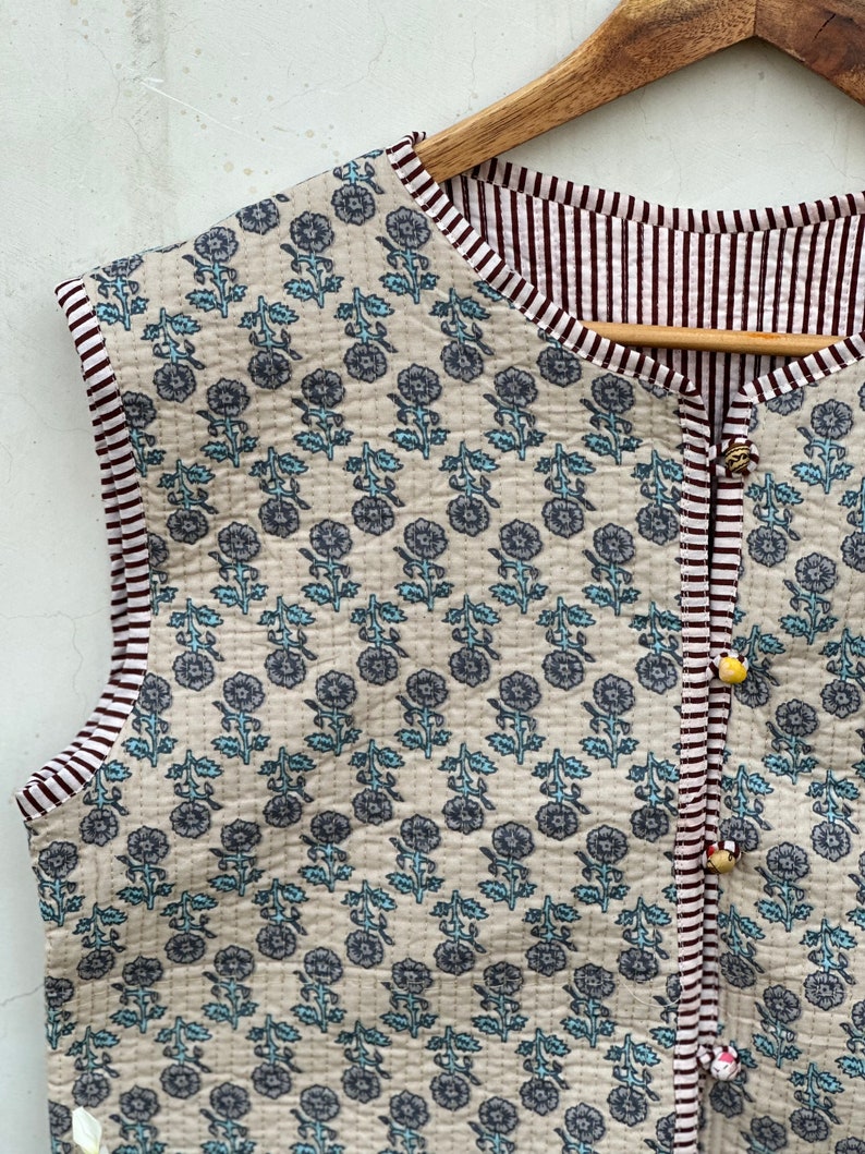 May include: A sleeveless, reversible quilted vest with a floral pattern in gray and light blue on a beige background. The vest has a striped border in white and burgundy, and is fastened with decorative buttons.