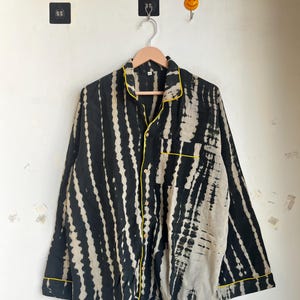 Może przedstawiać: Czarna i biała koszula z długim rękawem w stylu tie-dye z ż&oacute;łtymi lam&oacute;wkami. Koszula jest zapinana na guziki i ma kieszeń na piersi. Wisi na drewnianym wieszaku. Koszula ma unikalny, pionowy wz&oacute;r w paski.