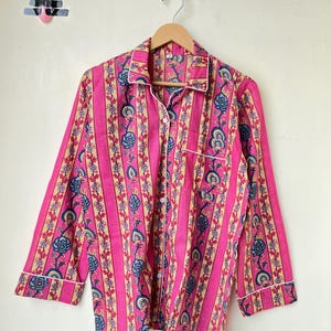 May include: Pink pajama top with vertical stripes and floral patterns in blue and beige. The long-sleeved shirt has a button-down front, a chest pocket, and white trim along the collar, cuffs, and pocket.