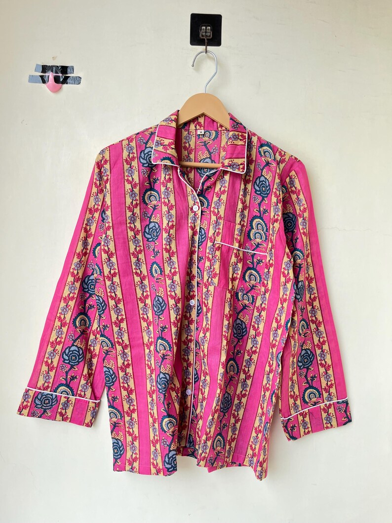 May include: Pink pajama top with vertical stripes and floral patterns in blue and beige. The long-sleeved shirt has a button-down front, a chest pocket, and white trim along the collar, cuffs, and pocket.