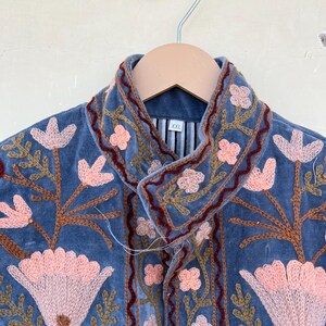 May include: A blue velvet jacket with embroidered floral details in shades of pink, brown, and green. The jacket has a collar and a button closure. The size tag reads "XXL".