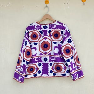 May include: A white jacket with a purple, blue, and orange geometric pattern embroidered on the front. The jacket has long sleeves and a round neckline.