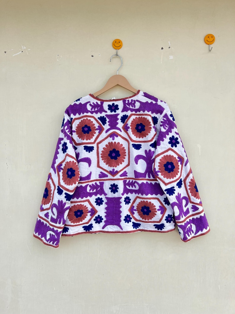 May include: A white jacket with a purple, blue, and orange geometric pattern embroidered on the front. The jacket has long sleeves and a round neckline.