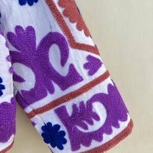 May include: White fabric with purple, blue, and pink embroidery. The embroidery is in a floral pattern.