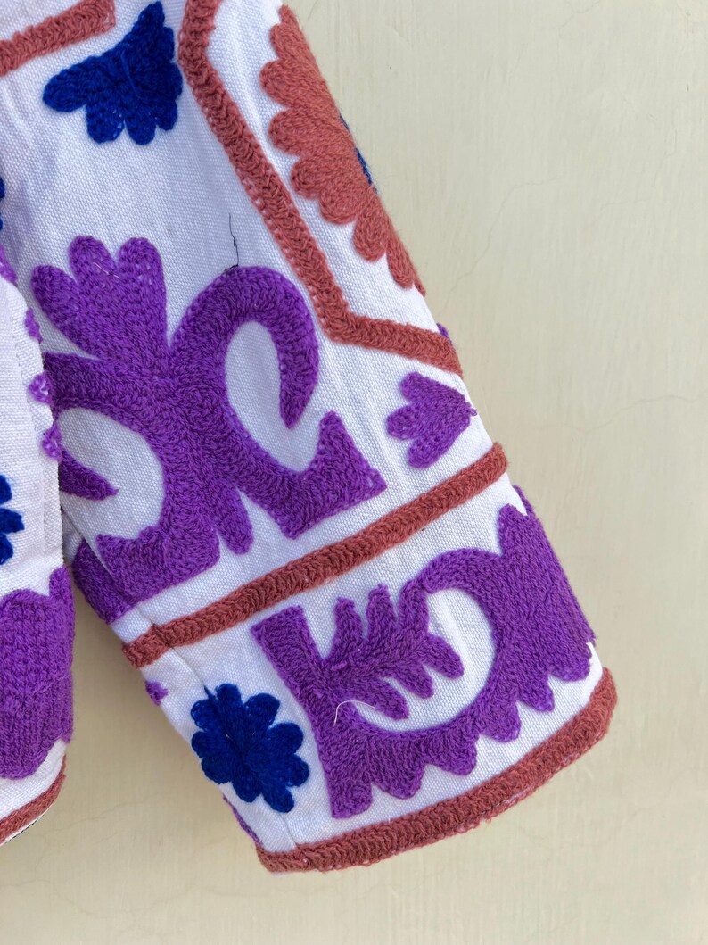 May include: White fabric with purple, blue, and pink embroidery. The embroidery is in a floral pattern.