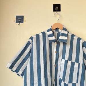 May include: A short-sleeved shirt with vertical stripes in white and blue. The shirt has a button-down front, a chest pocket, and wooden buttons. The shirt is hanging on a wooden hanger.