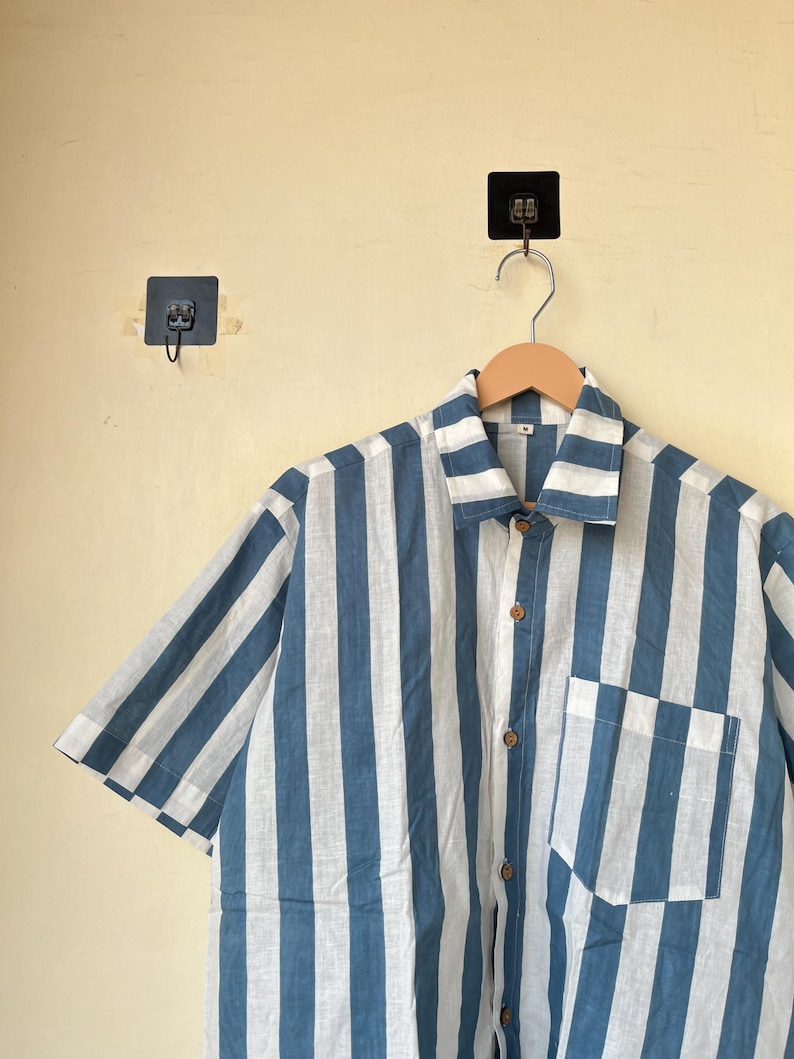 May include: A short-sleeved shirt with vertical stripes in white and blue. The shirt has a button-down front, a chest pocket, and wooden buttons. The shirt is hanging on a wooden hanger.