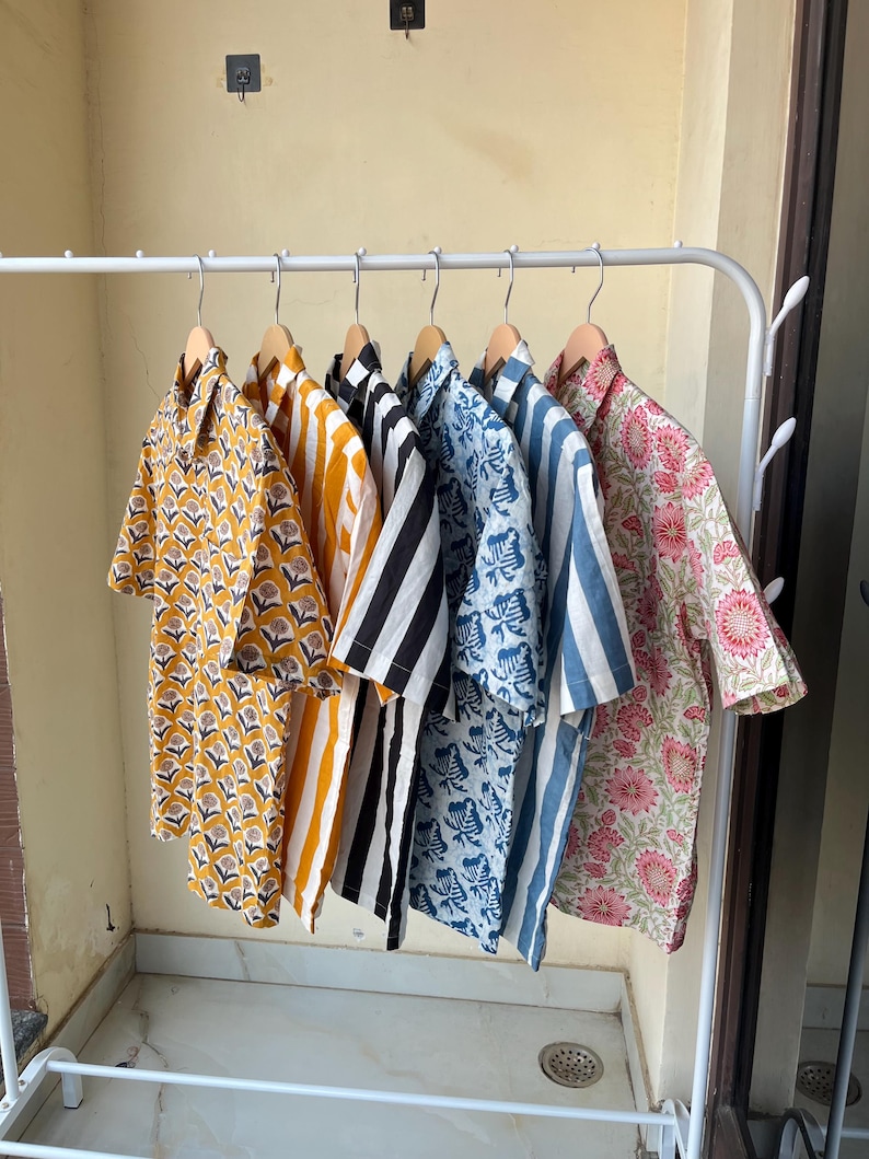 May include: A collection of short-sleeved shirts hanging on a white rack. The shirts feature various patterns, including floral, stripes, and geometric designs. Colors include yellow, black, white, blue, and pink. The shirts are made of a lightweight fabric.