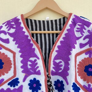 May include: White cotton jacket with purple, blue, and pink embroidery. The jacket has a V-neck and a tie closure.