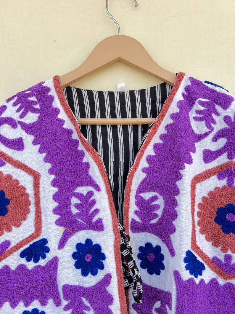 May include: White cotton jacket with purple, blue, and pink embroidery. The jacket has a V-neck and a tie closure.
