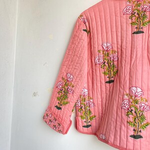 Pu&ograve; includere: Giacca trapuntata color corallo con stampa floreale. La giacca presenta un motivo ripetuto di fiori rosa e bianchi con steli verdi e basi nere. La giacca ha un colletto e maniche a tre quarti.