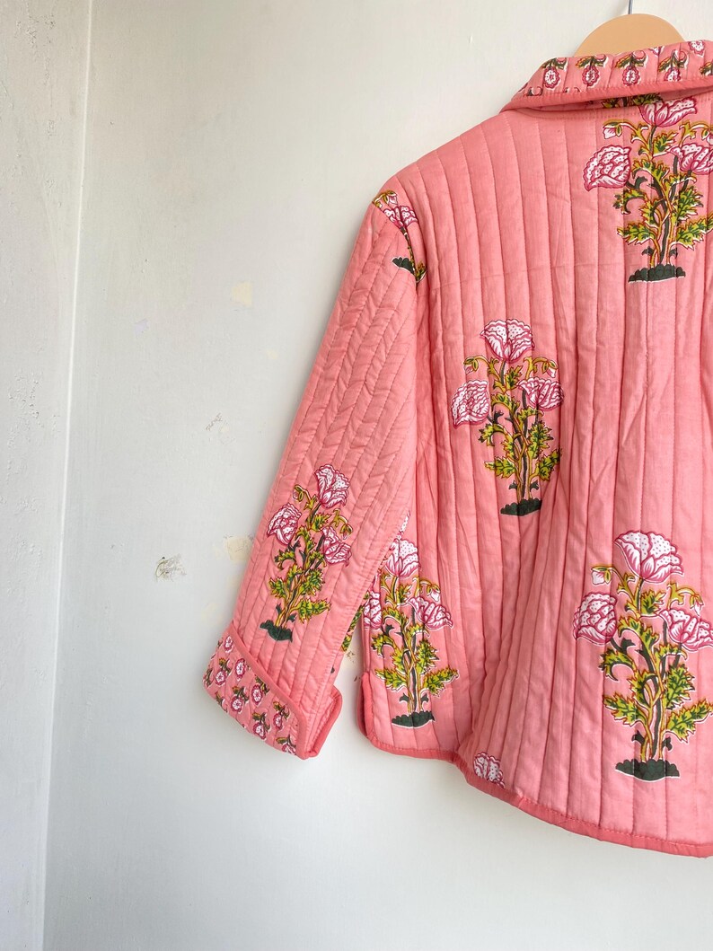 Pu&ograve; includere: Giacca trapuntata color corallo con stampa floreale. La giacca presenta un motivo ripetuto di fiori rosa e bianchi con steli verdi e basi nere. La giacca ha un colletto e maniche a tre quarti.